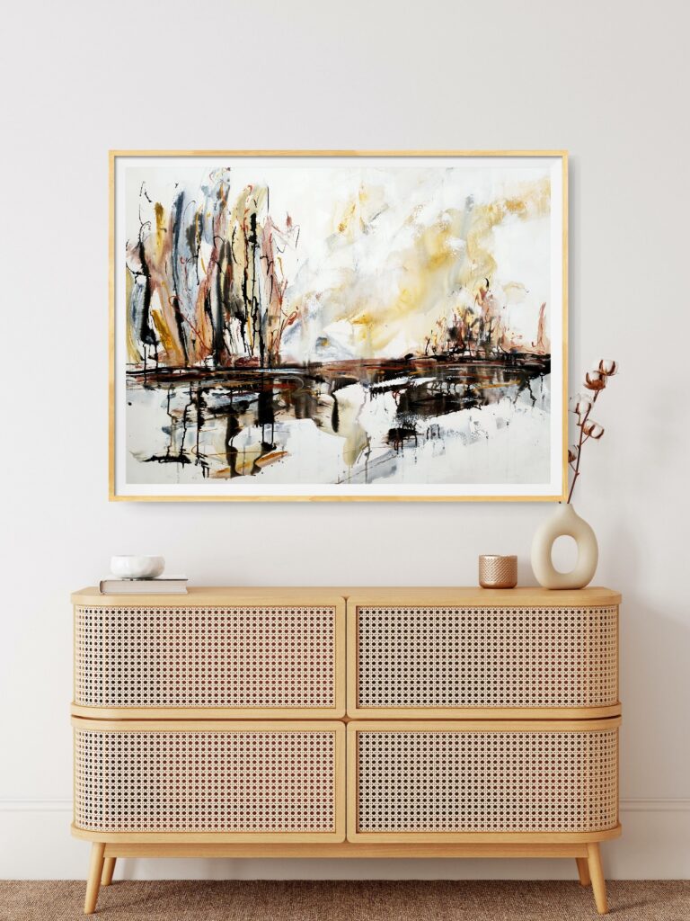 Lakeside Glory - An abstract landscape artwork portraying lake, trees, and the sky in natural colours of brown, ochre, black, white and grey. The painting is hanging on a pale wall, behind a wooden cabinet and decorating items.