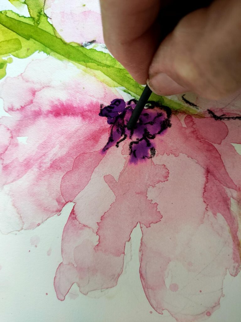 A mixed media painting of a pink flower and green stem showing part of a hand with a stick of charcoal drawing around the stamens.